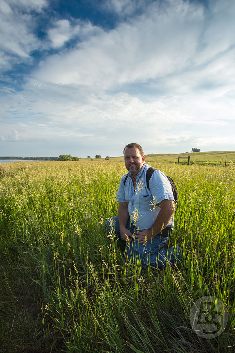 an image of the author in the early morning light among green prairie grass beneath a gorgeous blue sky with puffy white clouds