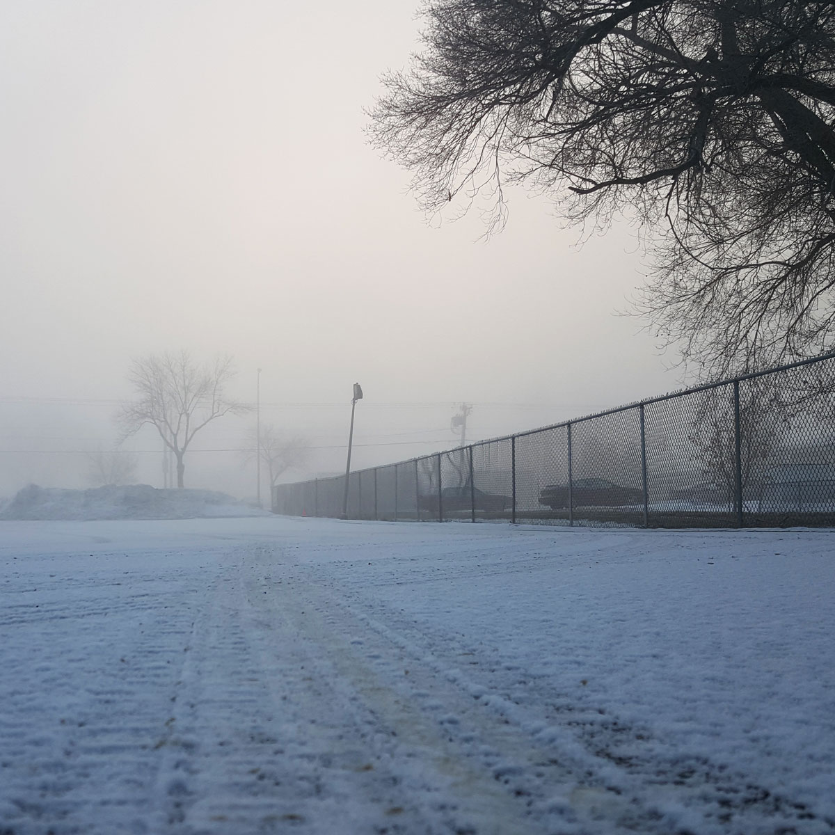 A foggy morning with a snow covered parking lot.In this low to the ground perspective shot a tire track in the snow leads into the distance.