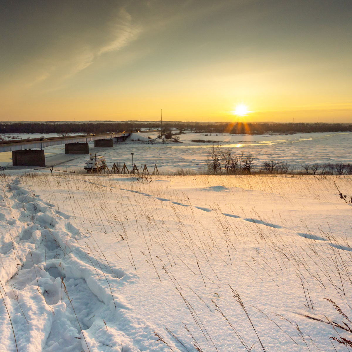 From a hilltop overlooking a frozen Missouri River and Keelboat Park in Bismarck, North Dakota a winter sunset brings golden colors to the sky and the reeds of grass that peek out above in the snow.