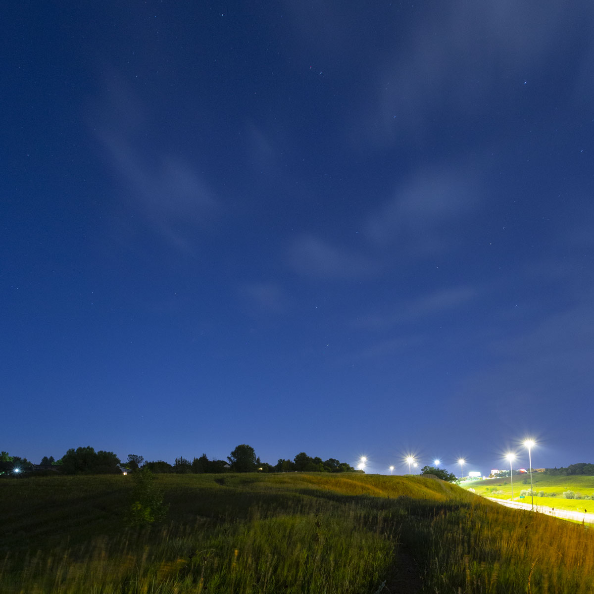 Through golden prairie grass in the dim light of dawn twilight a trail leads away toward a blue twilight sky filled with twinkling stars. The lights of a nearby interstate show as silvery starbursts.