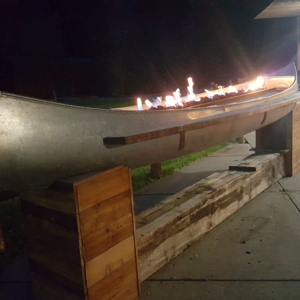 A nighttime image of an aluminum canoe sitting on wooden blocks  as a display on the porch at Spearfish Canyon Lodge. The interior of the canoe is lit stem to stern by flames dancing over lava rocks.