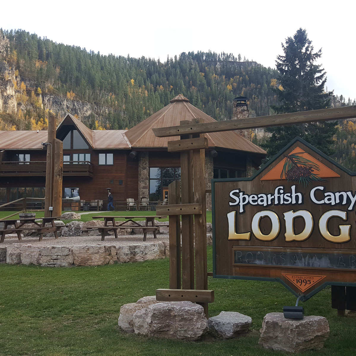 The warm, rustic wood exterior of Spearfish Canyon Lodge and the sign in front of it invites visitors in as Spearfish Canyon walls overlook in the distance.