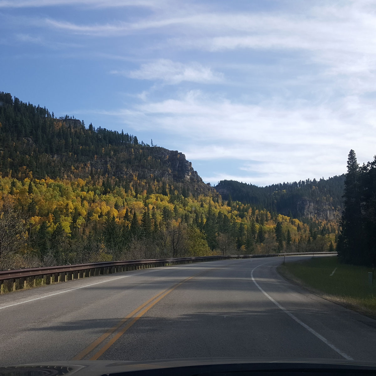 A windshield view of Spearfish Canyon Highway. A curve in the road leads right acccompanied by a guardrail while the beautiful Autumn colors in the canyon trees stand on the hills beyond.