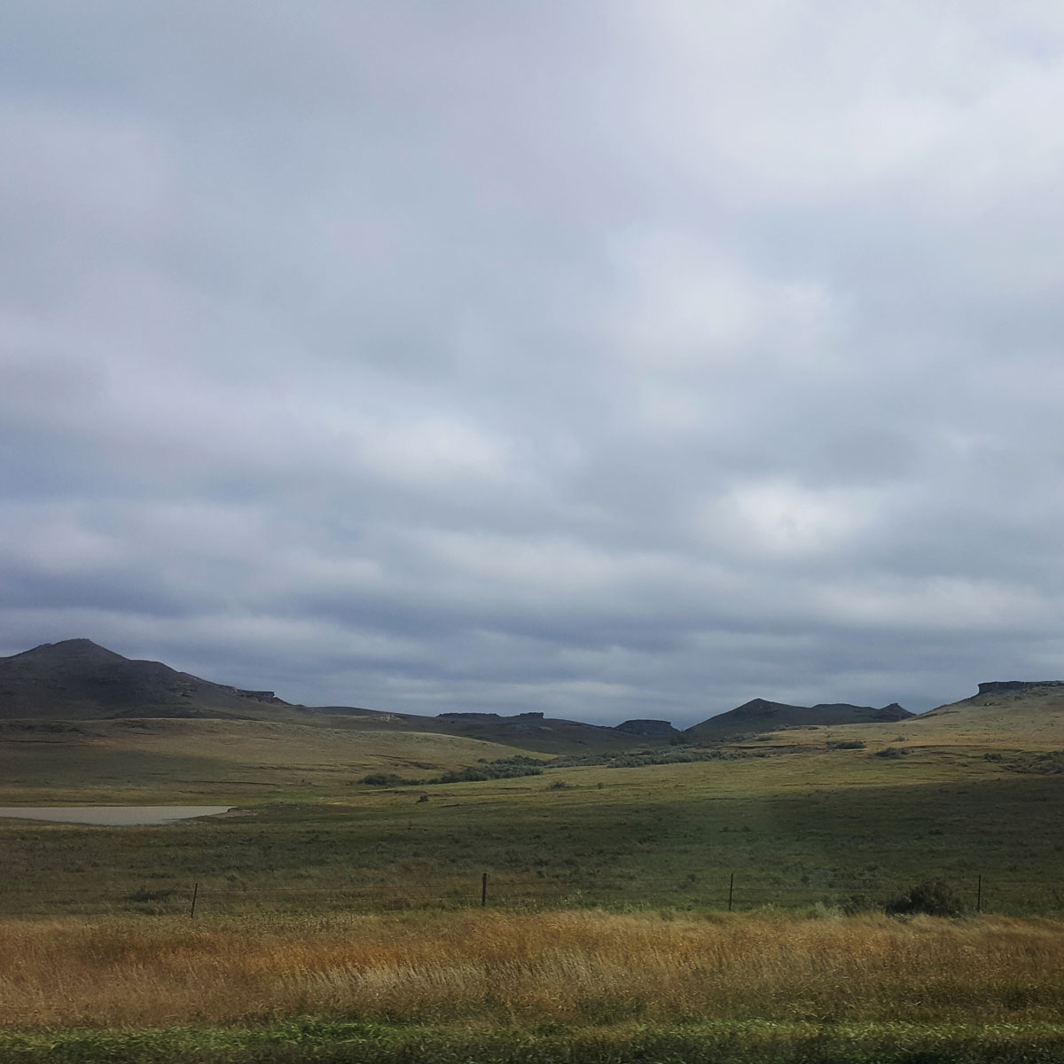 A view through a car window of buttes and hills under a cloudy sky in the distance across a grassy plain somewhere North of Belle Forche, South Dakota.