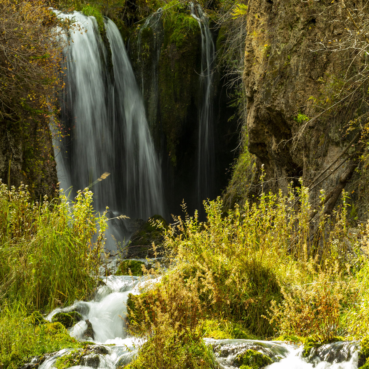 The main falls of Roughlock Falls in South Dakota spill down onto several smaller waterfalls surrounded by tall grasses backlit by golden late afternoon sunlit.